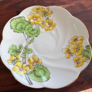 May include: A white porcelain saucer with a scalloped edge features a floral design of yellow flowers and green leaves. The saucer has a gold rim.