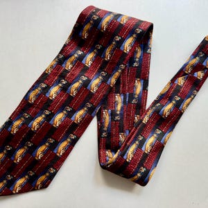 May include: A necktie featuring a repeating pattern of golf bags in tan and brown on a blue background, interspersed with vertical stripes of deep red and black. The fabric has a textured appearance with a pointed end and a narrow tail section.
