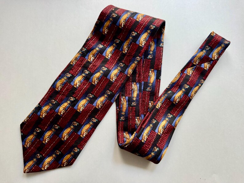 May include: A necktie featuring a repeating pattern of golf bags in tan and brown on a blue background, interspersed with vertical stripes of deep red and black. The fabric has a textured appearance with a pointed end and a narrow tail section.