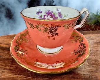 May include: A coral pink teacup and saucer set with a gold floral design and a painted violet flower motif.