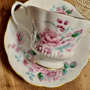 May include: A white teacup and saucer with a floral pattern in shades of pink, green, and blue. The cup has a scalloped edge and a gold trim.