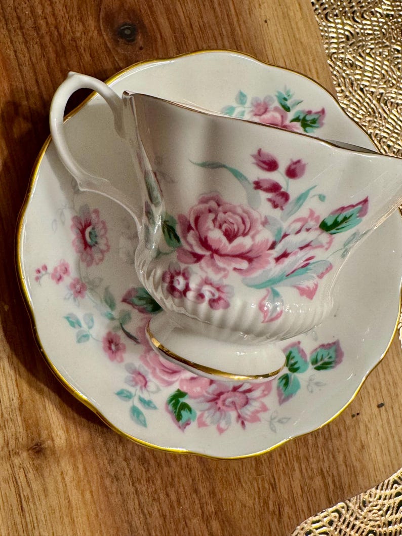 May include: A white teacup and saucer with a floral pattern in shades of pink, green, and blue. The cup has a scalloped edge and a gold trim.