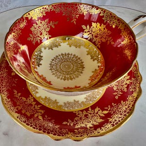 May include: A red teacup, saucer, and small plate set with gold floral detailing. The teacup has a white handle and a cream-colored interior. The saucer and plate have a scalloped edge.