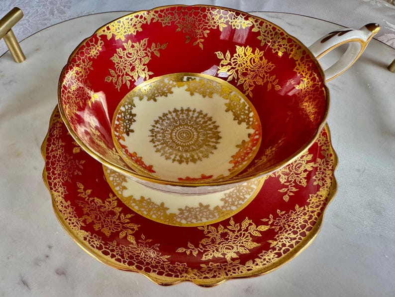 May include: A red teacup, saucer, and small plate set with gold floral detailing. The teacup has a white handle and a cream-colored interior. The saucer and plate have a scalloped edge.