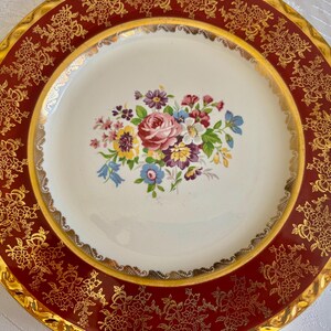 May include: A vintage decorative plate with a maroon border and gold floral filigree design. The center features a bouquet of pink, purple, blue, and yellow flowers on a white background. The plate has a gold rim.