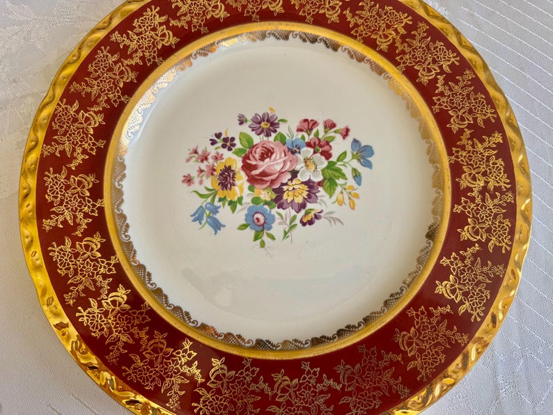 May include: A vintage decorative plate with a maroon border and gold floral filigree design. The center features a bouquet of pink, purple, blue, and yellow flowers on a white background. The plate has a gold rim.