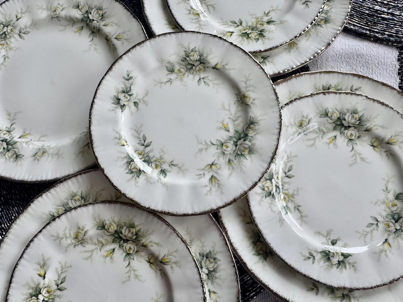 May include: A set of six white ceramic plates with a delicate floral design featuring white roses and green leaves. The plates have a scalloped edge and a gold rim.