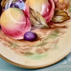 May include: A close-up of a decorative plate with a painted still life of fruit. The plate features peaches, grapes, and a plum. The plate has a gold rim and a cream-colored background.