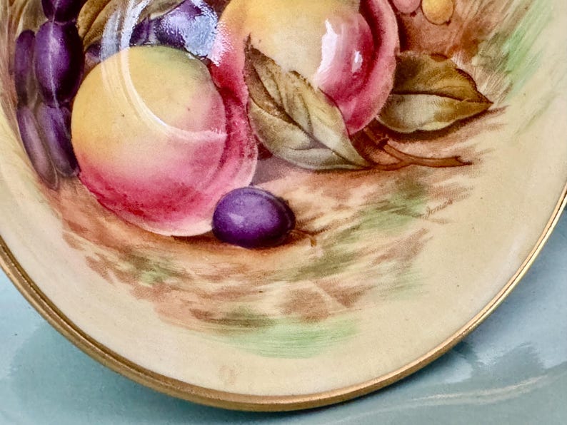 May include: A close-up of a decorative plate with a painted still life of fruit. The plate features peaches, grapes, and a plum. The plate has a gold rim and a cream-colored background.