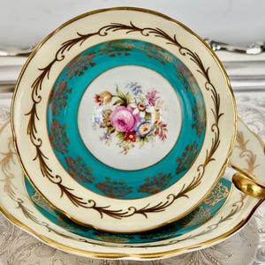 May include: A vintage teacup and saucer set. The teacup features a teal center with a floral bouquet and is bordered by a cream-colored rim with gold accents. The saucer mirrors the cup's design, with a gold-trimmed edge.