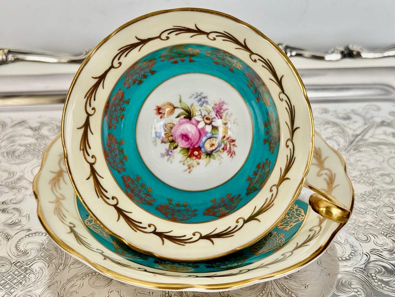 May include: A vintage teacup and saucer set. The teacup features a teal center with a floral bouquet and is bordered by a cream-colored rim with gold accents. The saucer mirrors the cup's design, with a gold-trimmed edge.