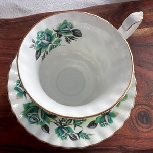 May include: A white teacup and saucer with a green rose pattern. The cup has a gold rim and the saucer has a scalloped edge.