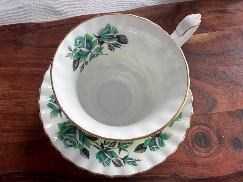 May include: A white teacup and saucer with a green rose pattern. The cup has a gold rim and the saucer has a scalloped edge.