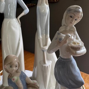 K&ouml;nnte beinhalten: Eine Sammlung von Porzellanfiguren. Die Figuren zeigen Frauen und M&auml;dchen in verschiedenen Posen, mit sanften Pastellfarben. Eine Figur h&auml;lt einen Vogel und einen Korb. Die Figuren stehen auf einem marmor&auml;hnlichen Sockel.
