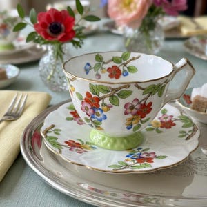K&ouml;nnte beinhalten: Wei&szlig;es Teeservice mit floralem Muster. Die Tasse und Untertasse haben einen Goldrand und einen hellgr&uuml;nen Sockel. Das florale Muster umfasst rote, blaue und gelbe Blumen. Das Set steht auf einem silbernen Tablett.