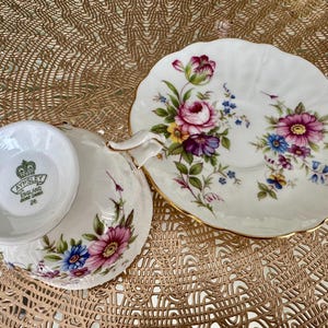 May include: A white teacup and saucer with a floral pattern. The saucer has a gold rim and the teacup has a gold band around the top. The teacup has the words "Aysnley England Bone China 26" on the bottom.