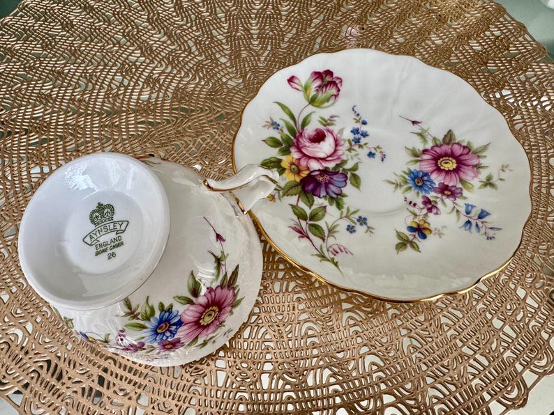 May include: A white teacup and saucer with a floral pattern. The saucer has a gold rim and the teacup has a gold band around the top. The teacup has the words "Aysnley England Bone China 26" on the bottom.