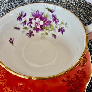 May include: A red teacup and saucer with a gold rim and a floral design featuring purple and white flowers. The saucer has a gold floral pattern.