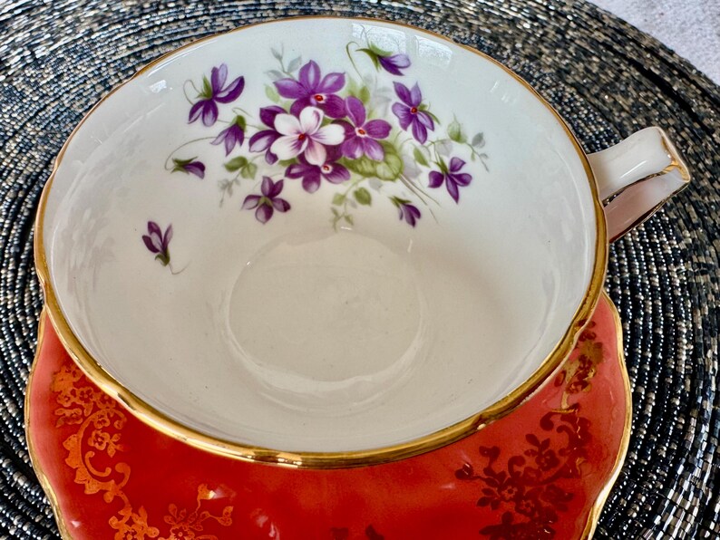 May include: A red teacup and saucer with a gold rim and a floral design featuring purple and white flowers. The saucer has a gold floral pattern.