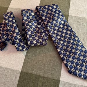 May include: A necktie featuring a repeating checkered pattern of dark blue squares and white soccer balls. The tie has a pointed end and a folded structure, displaying a consistent graphic print of black and white soccer balls throughout the fabric.