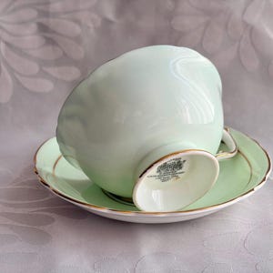 May include: A pale mint green porcelain teacup and saucer set featuring a scalloped edge design and delicate gold trim along the rim and base. The underside of the teacup displays a black maker's mark with text including Bone China.