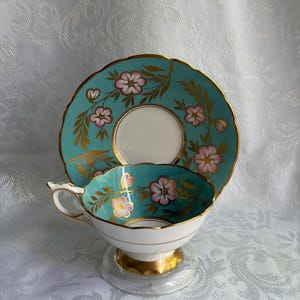 May include: A porcelain teacup and saucer set featuring a vibrant turquoise background with hand-painted pink flowers and gold leaf accents. The cup has a scalloped rim and a gold-trimmed handle, while the saucer displays a matching floral pattern.