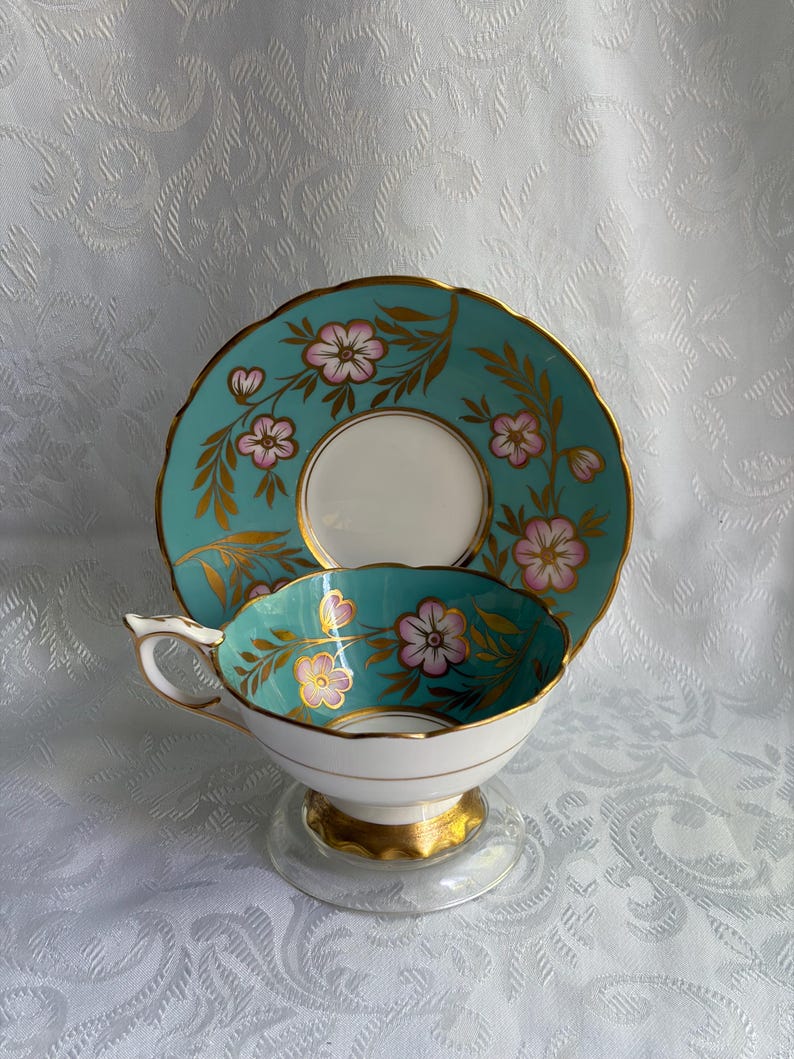 May include: A porcelain teacup and saucer set featuring a vibrant turquoise background with hand-painted pink flowers and gold leaf accents. The cup has a scalloped rim and a gold-trimmed handle, while the saucer displays a matching floral pattern.