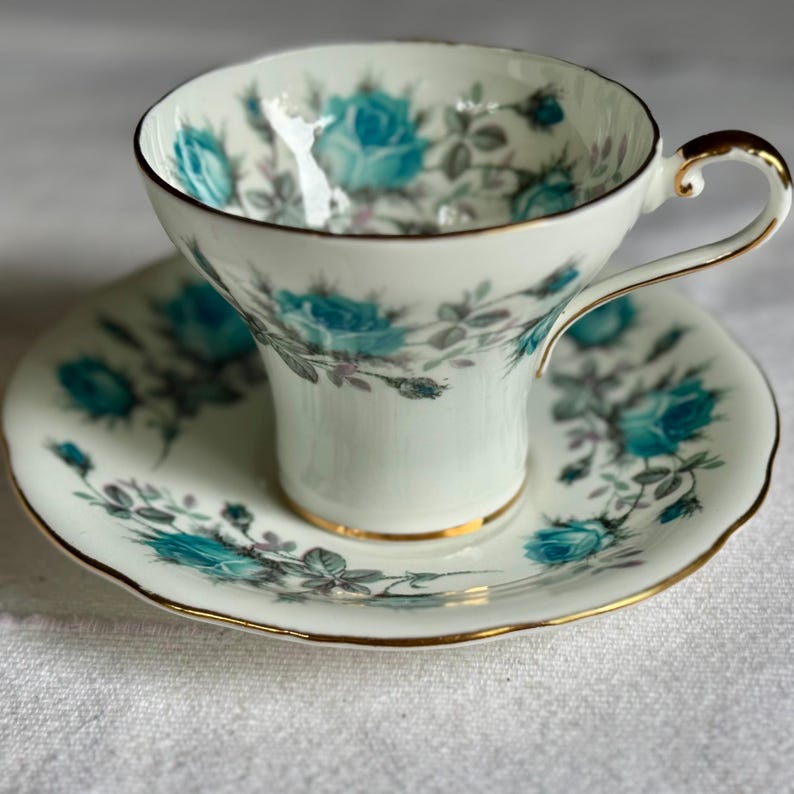 Peut inclure: Une tasse et une soucoupe blanches avec un motif de roses turquoise et une bordure dor&eacute;e. La tasse a une anse incurv&eacute;e et repose sur une soucoupe assortie. Le motif floral comprend des roses turquoise et des feuilles grises.