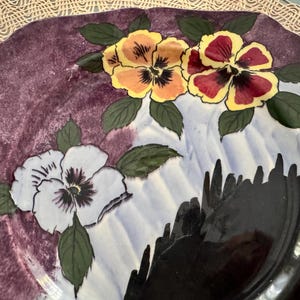 May include: A decorative ceramic plate with a floral design. The plate features three pansies in shades of yellow, red, and white, with green leaves. The background is a mix of purple and white, with a black rim.
