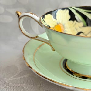 May include: A pale mint green teacup and saucer set featuring a gold-trimmed handle and rim. The interior of the cup displays a black background with a large yellow flower pattern. The saucer has a matching mint green finish with gold accents.