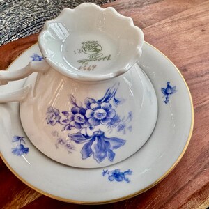 May include: A white teacup and saucer with a blue floral pattern. The teacup has a scalloped edge and the saucer has a gold rim. The bottom of the teacup has a green and gold mark that reads "Royal Copenhagen 1964".