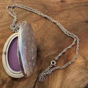 Puede incluir: Collar con medall&oacute;n ovalado plateado con tapa abierta que revela un interior morado. El medall&oacute;n tiene un dise&ntilde;o floral grabado y cuelga de una delicada cadena. El collar descansa sobre una superficie de madera.