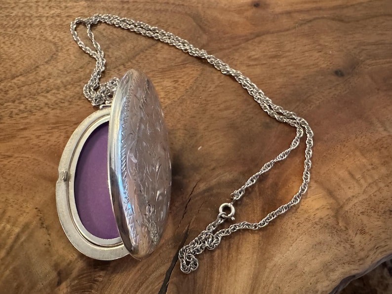 Puede incluir: Collar con medall&oacute;n ovalado plateado con tapa abierta que revela un interior morado. El medall&oacute;n tiene un dise&ntilde;o floral grabado y cuelga de una delicada cadena. El collar descansa sobre una superficie de madera.