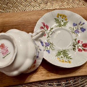 May include: A white teacup and saucer with a floral pattern. The saucer has a scalloped edge and features a variety of flowers, including pink, blue, and yellow. The teacup has a floral pattern on the outside and the words "Paragon English Bone China" on the bottom.