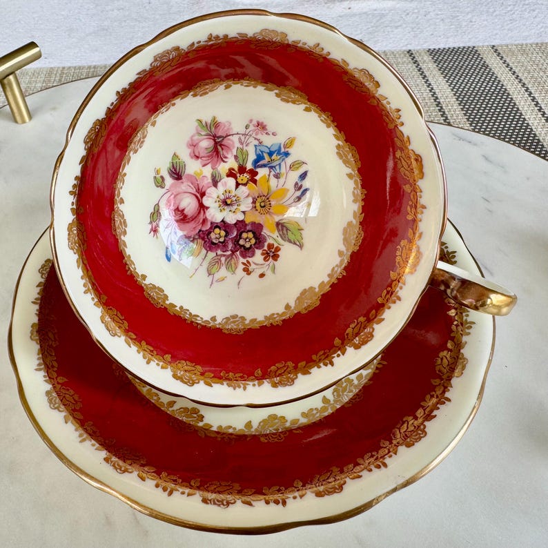 May include: A red and gold teacup and saucer set with a floral design. The cup and saucer have a scalloped edge and a gold trim.