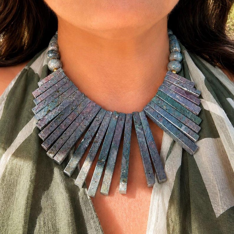 May include: A statement necklace featuring a series of rectangular, vertically oriented stone bars in shades of mottled green, blue, and purple. The bars are suspended from a strand of round, speckled grey beads that connect at the back of the neck.