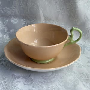 May include: A peach-colored teacup and saucer set featuring a scalloped rim design. The teacup has a light green handle and a matching light green base, while the saucer is peach-colored with a white underside, creating a two-tone appearance.