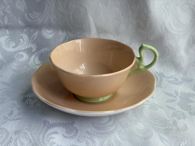May include: A peach-colored teacup and saucer set featuring a scalloped rim design. The teacup has a light green handle and a matching light green base, while the saucer is peach-colored with a white underside, creating a two-tone appearance.
