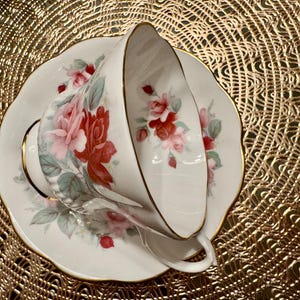 May include: A white teacup and saucer with a floral pattern of pink and red roses. The saucer has a gold trim.