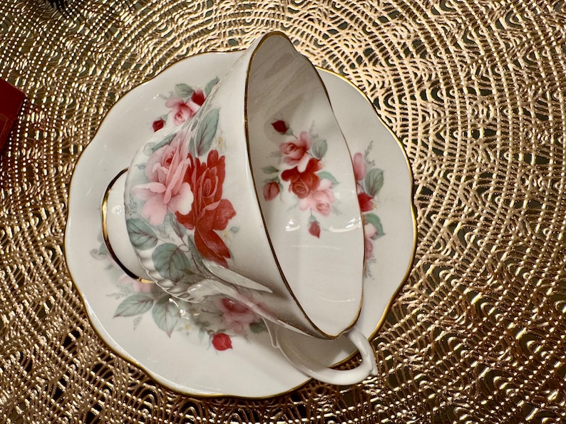 May include: A white teacup and saucer with a floral pattern of pink and red roses. The saucer has a gold trim.