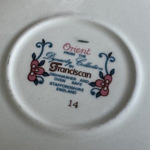 Może przedstawiać: Biały ceramiczny talerz z napisem "Orient from the Dynasty Collection by Franciscan" w kolorze turkusowym i r&oacute;żowym. Talerz ma r&oacute;wnież kwiatowy wz&oacute;r i numer "14". Można myć w zmywarce i piekarniku.