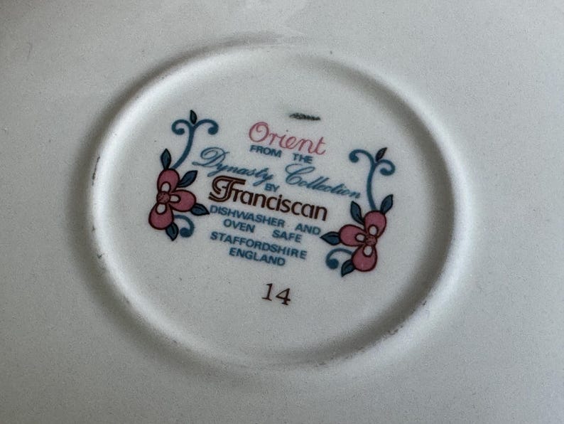 Może przedstawiać: Biały ceramiczny talerz z napisem "Orient from the Dynasty Collection by Franciscan" w kolorze turkusowym i r&oacute;żowym. Talerz ma r&oacute;wnież kwiatowy wz&oacute;r i numer "14". Można myć w zmywarce i piekarniku.