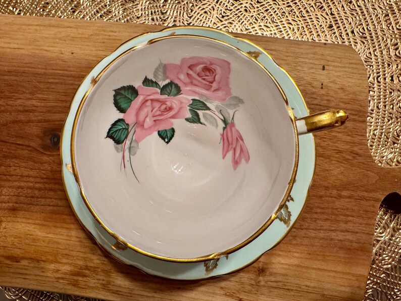 May include: A light blue teacup and saucer with a gold rim. The teacup features a floral design with pink roses and green leaves.