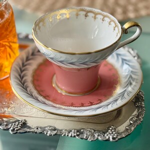 May include: A vintage teacup and saucer set with a pink base and gold trim, resting on a silver tray. The cup has a white interior and a delicate gold pattern along the rim. The saucer features a matching pink center and a detailed design.