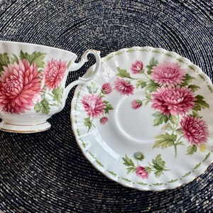 May include: A white teacup and saucer set with a floral design. The teacup and saucer are decorated with pink and red flowers and green leaves. The edges of the teacup and saucer are trimmed with gold.