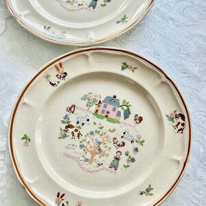 May include: Two off-white ceramic plates with a brown rim and scalloped edges. Each plate features a pastoral scene with a pink house, trees, animals, and a woman in a dress. Floral accents are scattered around the edges.