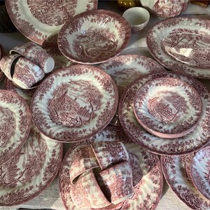 Pu&ograve; includere: Una collezione di stoviglie antiche bianche e bordeaux, tra cui piatti, ciotole e tazze. I piatti presentano un design paesaggistico dettagliato con un bordo floreale. Il set include piatti, ciotole e tazze di varie dimensioni.