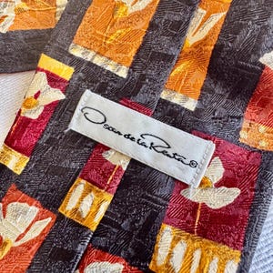 Op de afbeelding: Close-up van een Oscar de la Renta stropdas. De stropdas heeft een herhalend patroon van bloemmotieven in oranje, rood en wit tegen een donkere achtergrond. Het label "Oscar de la Renta" is zichtbaar.