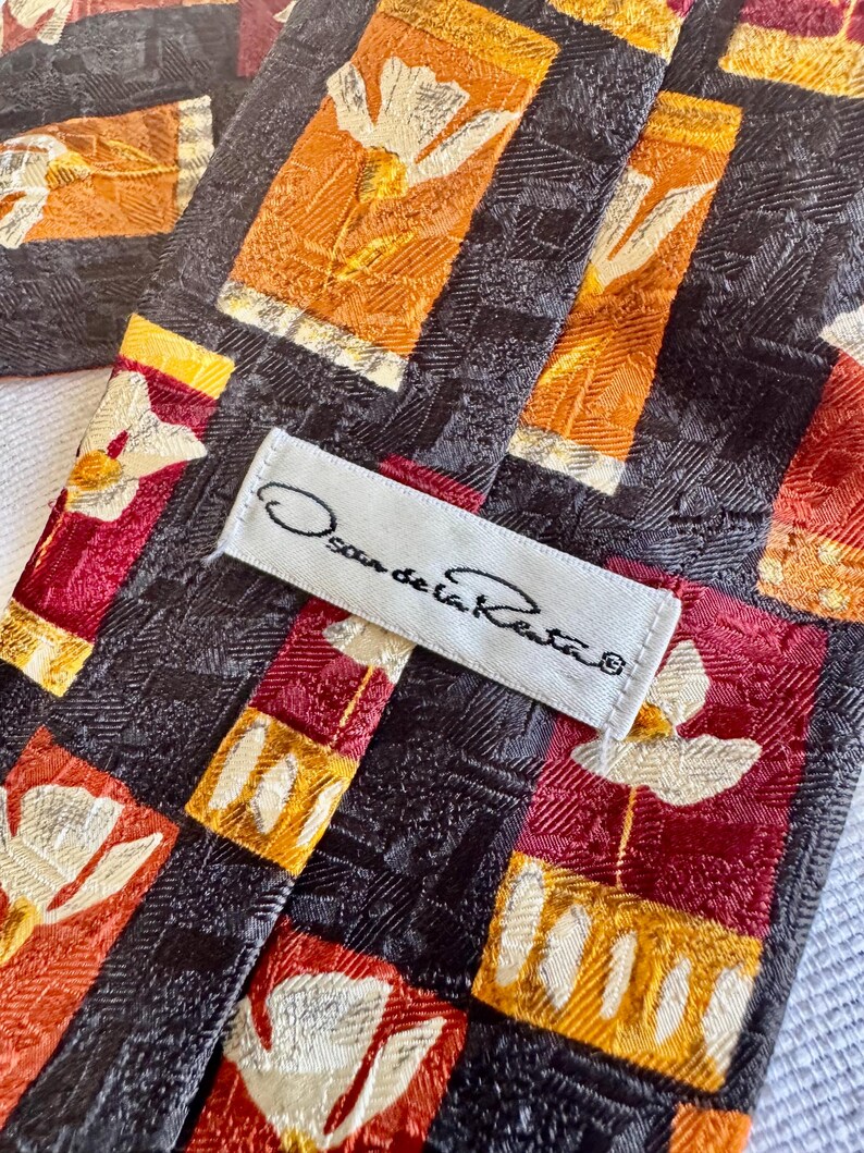 Op de afbeelding: Close-up van een Oscar de la Renta stropdas. De stropdas heeft een herhalend patroon van bloemmotieven in oranje, rood en wit tegen een donkere achtergrond. Het label "Oscar de la Renta" is zichtbaar.