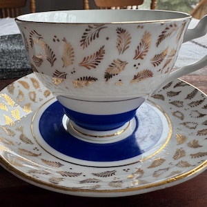May include: A white teacup and saucer with a gold leaf pattern and a blue band around the base of the cup. The saucer has a gold leaf pattern.